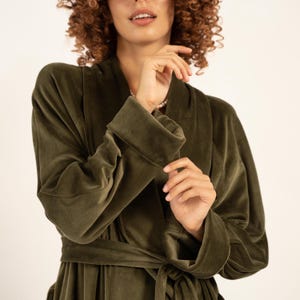May include: An olive green velvet dressing gown with a shawl collar and a matching belt. The gown has long sleeves with folded cuffs. The model is wearing a pearl necklace. The gown is made of a soft, plush material.