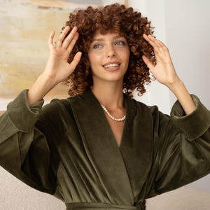 May include: An olive green velvet robe with wide sleeves and a wrap-around design. The robe is worn with a pearl necklace. The model has curly brown hair.