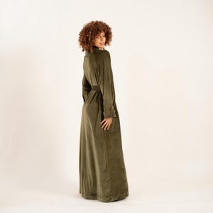 May include: An olive green velvet robe with a matching belt. The robe has long sleeves and a high collar. The model is wearing the robe and is turned to the side, against a neutral background.