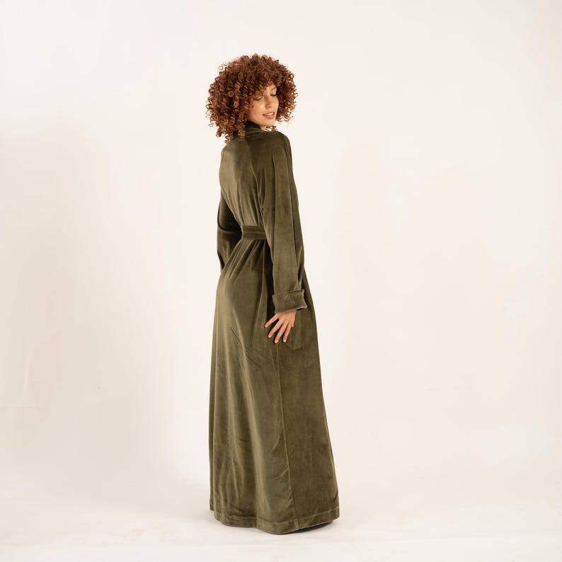 May include: An olive green velvet robe with a matching belt. The robe has long sleeves and a high collar. The model is wearing the robe and is turned to the side, against a neutral background.