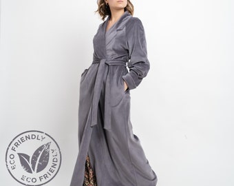 Puede incluir: Una bata larga de color gris con cuello chal y cinturón de lazo. La bata tiene bolsillos a los lados y una larga abertura en la parte delantera. La bata está hecha de una tela suave y afelpada. La etiqueta de la bata dice "ECO FRIENDLY".