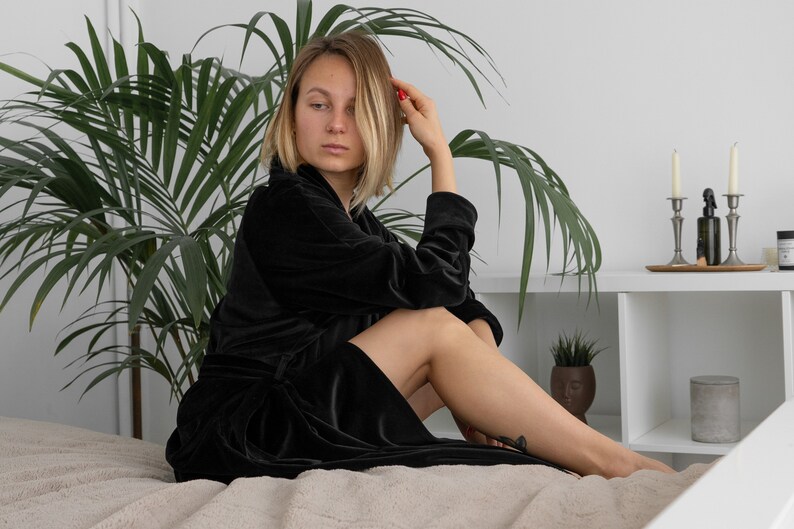 May include: A woman wearing a black velvet robe sits on a bed with a plant in the background.