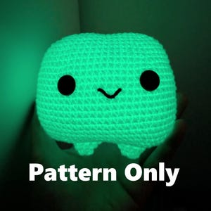 Op de afbeelding: Een gloeiend groen gehaakt wezen met zwarte ogen en een lachende mond. Het handgemaakte pluche speelgoed heeft een rechthoekig lichaam en kleine voetjes. De tekst "Pattern Only" is zichtbaar onderaan de afbeelding.