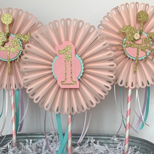 May include: Three pink paper fans with gold glitter accents and a carousel horse design. The center fan has a pink number one with gold glitter. The fans are attached to sticks with pink and white striped paper straws and pink, white, and teal ribbons.