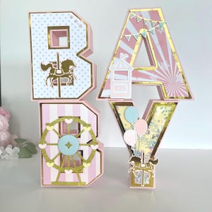 May include: Pink and gold decorative letters spelling "BABY". The letters feature carnival-themed designs, including a carousel horse, Ferris wheel, and balloons. The letter "A" has a banner of small flags.