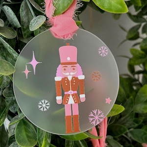 Könnte beinhalten: Ein klarer, runder Weihnachtsschmuck mit einem rosa-braunen Nussknackersoldaten-Design. Der Schmuck hat rosa Stern- und Schneeflocken-Akzente und wird von einem rosa Band gehalten. Der Schmuck ist von grünem Laub umgeben.