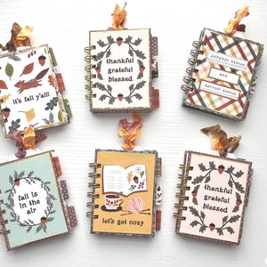 Könnte beinhalten: Sechs spiralgebundene Mini-Notizbücher mit herbstlichen Motiven, verziert mit Blättern, Eicheln und Texten wie "it's fall y'all", "thankful grateful blessed", "pumpkin kisses and harvest wishes", "fall is in the air" und "let's get cozy".