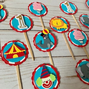 May include: A collection of circus-themed cupcake toppers. Each topper features a different design, including a lion, elephant, clown, ice cream cone, and a circus tent. The toppers are made of layered paper with a blue and red color scheme.