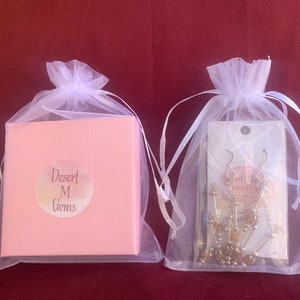May include: Two white organza bags, one containing a pink box with the text "Desert M Gems" and the other containing a pair of gold dangle earrings.