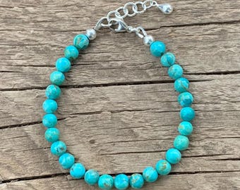 May include: A turquoise beaded bracelet with a silver-colored clasp and adjustable chain. The round beads are a vibrant blue-green color, strung together to form a complete circle. The bracelet is displayed on a rustic wooden surface.