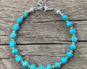 May include: A turquoise beaded bracelet with silver-colored accents. The bracelet features round, light blue beads separated by small silver beads. The adjustable chain and clasp are also silver. The bracelet is displayed on a wooden surface.