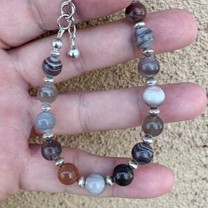 May include: A bracelet made with agate beads in shades of brown, gray, and white. The beads are separated by silver spacer beads. The bracelet has a silver clasp.