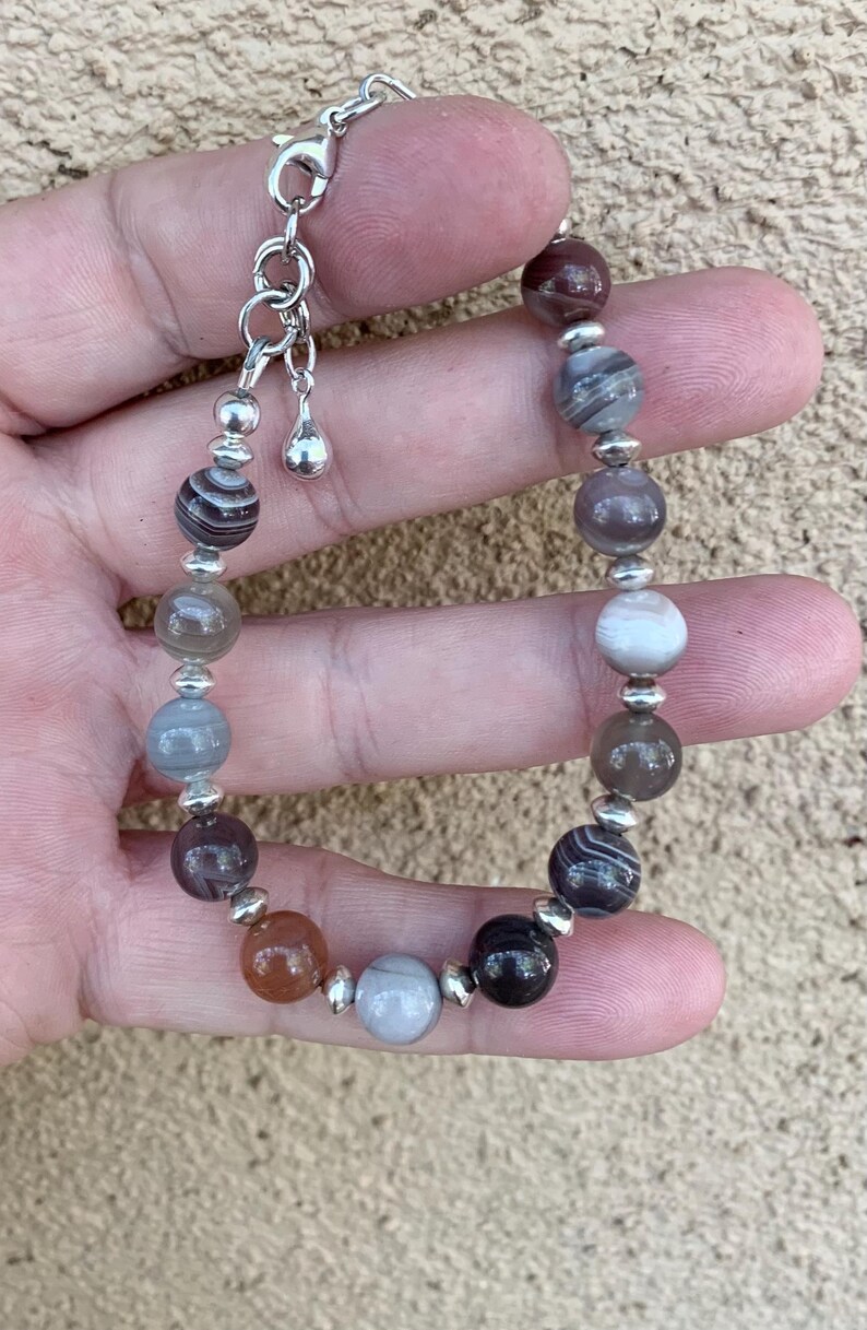May include: A bracelet made with agate beads in shades of brown, gray, and white. The beads are separated by silver spacer beads. The bracelet has a silver clasp.