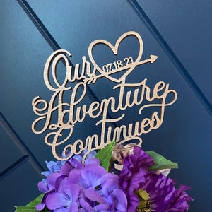 May include: Wooden cake topper with the words "Our Adventure Continues" and a heart with the date "07.18.21" inside. The topper is decorated with purple flowers.