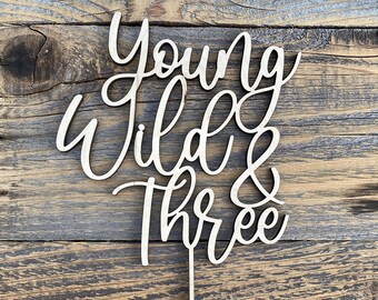 May include: Wooden cake topper with the words "Young Wild & Three" in a cursive font.