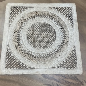 May include: Square, handmade placemat with a woven design. The center features a circular pattern with concentric rings and a woven texture in shades of brown and white. The border is a solid white.