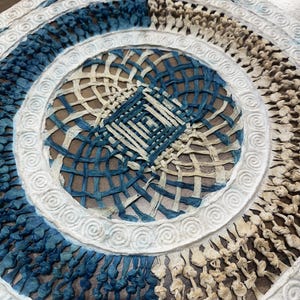 May include: Close-up of a woven decorative piece with a circular design. The piece features a combination of blue, off-white, and beige tones. The design includes concentric circles and a central square, all with intricate woven patterns and a textured surface.