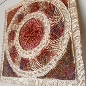 May include: A square wall art piece featuring a central circular woven design in shades of red, orange, and purple. The border consists of a raised, textured cream-colored frame with a repeating spiral pattern surrounding the woven center.