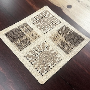 May include: A square, decorative placemat with a cream-colored border and four interior squares. Two squares feature a woven pattern in shades of brown, while the other two display a spiral and wavy design in beige. The border has a repeating swirl pattern.