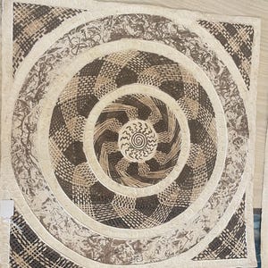 May include: A square, off-white, woven rug with a circular pattern in shades of brown. The center of the rug features a spiral design with a sunburst pattern.
