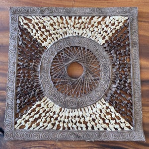 May include: Square, handmade wall art with a woven design. The artwork features a central circular pattern and a square frame with a repeating spiral design. The colors are neutral, with shades of brown and cream.
