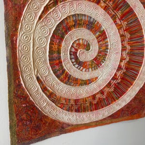 May include: A square wall art piece featuring a raised, white, spiral-patterned relief centered on a textured, multi-colored background of deep reds, oranges, and browns. The spiral contains intricate, thin, vertical lines in warm earth tones.