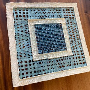 May include: A square, handmade paper placemat with a layered design. The outer layer is white with a textured edge, framing a light blue, patterned layer. The center features a woven blue square with a white, decorative border.