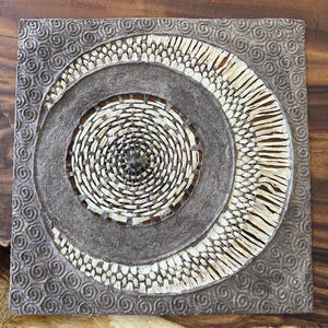 May include: Square, textured wall art with a circular woven design in the center. The artwork features a spiral pattern in the middle, surrounded by a dark brown ring and a crescent-shaped woven border. The outer border has a repeating swirl pattern.