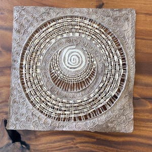 May include: A square, tan-colored woven placemat with a circular, spiral design in the center. The outer border features a repeating swirl pattern. The inner circle is made of woven natural fibers.