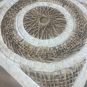 May include: A square, woven wall art piece with a spiral design. The center features a radial pattern, surrounded by concentric circles of woven material. A white, textured border with spiral details frames the artwork.