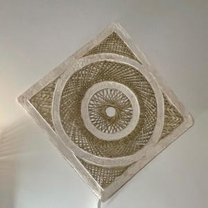 May include: A woven, square-shaped light fixture with a circular design in the center. The fixture is made of natural-colored woven material and has a white border. The design features concentric circles and a diamond shape.