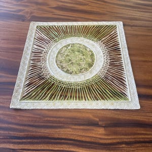 May include: A square, off-white decorative piece with a circular design. The center features a textured, light green material, surrounded by concentric circles and radiating lines. The border has a repeating swirl pattern, all set against a dark wood surface.