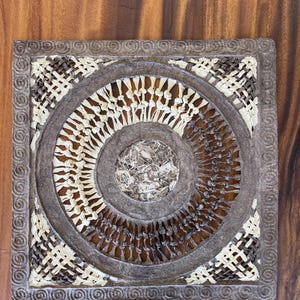 May include: Square, handcrafted wall art with a circular design. The artwork features a woven pattern in shades of brown and cream, with a textured, layered appearance. The outer frame has a decorative border.