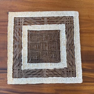May include: Square coaster with a woven brown center, surrounded by a cream-colored border with a spiral design. The coaster has a natural, handmade appearance, suitable for home decor.