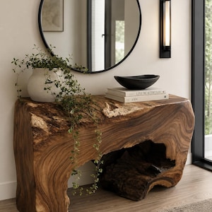 May include: A live-edge wooden console table with a natural, organic shape. A black bowl and books are on top, with a white vase holding greenery. A black-framed mirror and wall sconce are in the background. The table is approximately 60 inches long.