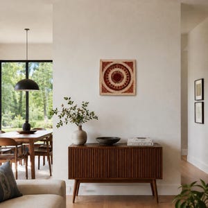 May include: A square wall art piece featuring concentric circles in shades of deep red and cream, mounted on a light-colored wall above a dark brown wooden sideboard with vertical slats, a ceramic vase with green branches, and a dark bowl.