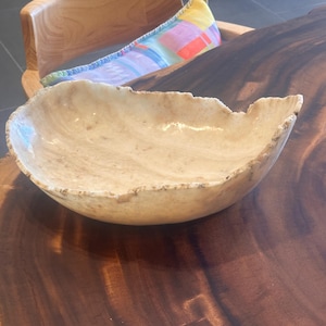 May include: A handcrafted, beige-colored bowl with a unique, organic shape. The bowl has a rough, textured edge and a smooth interior. The bowl is sitting on a dark brown wooden surface. A wooden chair and a colorful pillow are in the background.