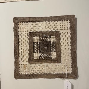 May include: A square wall hanging made of woven paper with a brown border and a white center. The center has a smaller square with a woven pattern in brown and white. The hanging has a price tag that says "49.99".