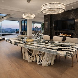 May include: A modern dining room featuring a large, rectangular table with a unique, natural wood base and a black and white patterned stone top. The room includes a fireplace, a large TV, and a staircase. Two crystal chandeliers hang from the ceiling.