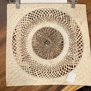 May include: A square, cream-colored paper wall hanging with a circular, woven design in the center. The woven design is made of thin strips of paper that are interlaced to create a pattern of concentric circles. The outer edge of the wall hanging is decorated with a repeating pattern of small, swirling circles.