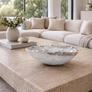May include: A white and gray decorative bowl with a textured rim sits on a light brown rectangular coffee table. The bowl is the focal point, with a neutral-toned living room in the background. A vase with flowers and a book are also on the table.
