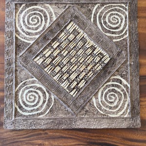 May include: Square, brown decorative wall art with a woven center and white spiral designs. The artwork has a textured border and is set against a wooden surface. The woven center is a light tan color.