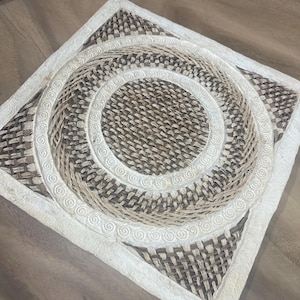 May include: Square woven placemat with a central spiral design. The placemat features a natural color palette with a woven texture. The border is white with a repeating swirl pattern. The placemat is designed for dining and home decor.