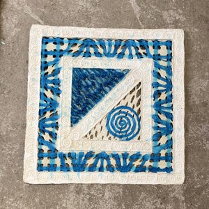May include: A white square cushion with a blue and white geometric pattern. The cushion has a white border with a blue and white swirl design.