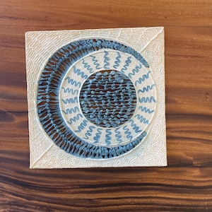 May include: Square, off-white artwork with a circular design. The design features a blue crescent shape and a central circle filled with small blue elements. The background has a textured pattern of swirls and lines.