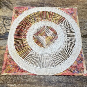 May include: A square, white, woven wall hanging with a circular design in the center. The center circle has a smaller square design inside of it. The woven design is made of natural fibers and has a light brown, yellow, and red color scheme.