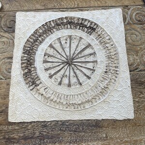May include: A white square wall hanging with a brown circular design in the center. The design is made of woven paper and has a radial pattern. The background of the wall hanging is textured with small white circles.