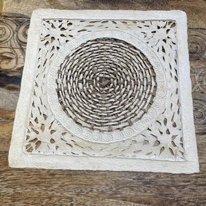 May include: A white square coaster with a woven circular design in the center. The coaster has a decorative, intricate pattern around the woven circle.
