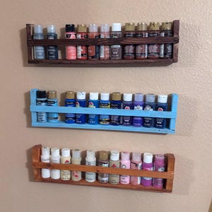 May include: Three wooden wall shelves with rows of small paint bottles. The top shelf is dark brown, the middle shelf is light blue, and the bottom shelf is light brown. The paint bottles are various colours, including red, orange, yellow, green, blue, purple, pink, and white.