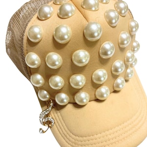 May include: A tan baseball cap featuring a mesh back and embellished with multiple faux pearls. A gold-coloured treble clef charm, set with rhinestones, dangles from the brim. The cap combines casual and elegant design elements.